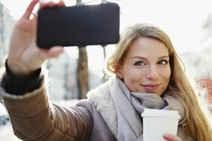 pohľadná žena, ktorá sa v zime prechádza ulicami mesta a robí si selfie a drží kávu so sebou, podsvietená