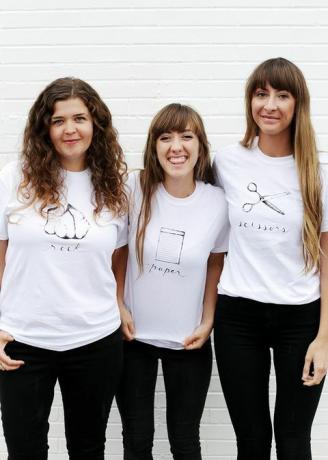 tri ženy v bielych tričkách, ktoré majú na košeli napísané a zobrazené buď kamene, papier alebo nožnice