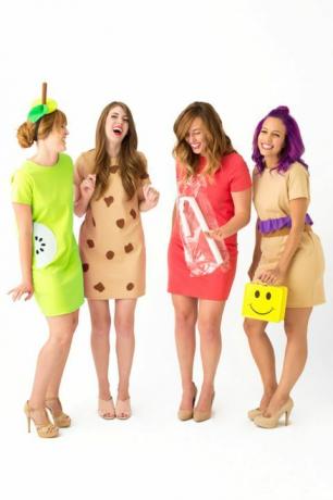 štyri vysmiate ženy v krátkych šatách oblečené ako „obedové dámy“, jedna nesúca obedovú krabičku so žltým smajlíkom